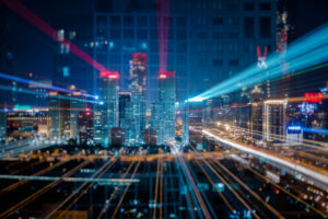 Paisagem urbana noturna com arranha-céus iluminados e trilhas de luz representando dados e conectividade digital, simbolizando a infraestrutura tecnológica de uma cidade inteligente.